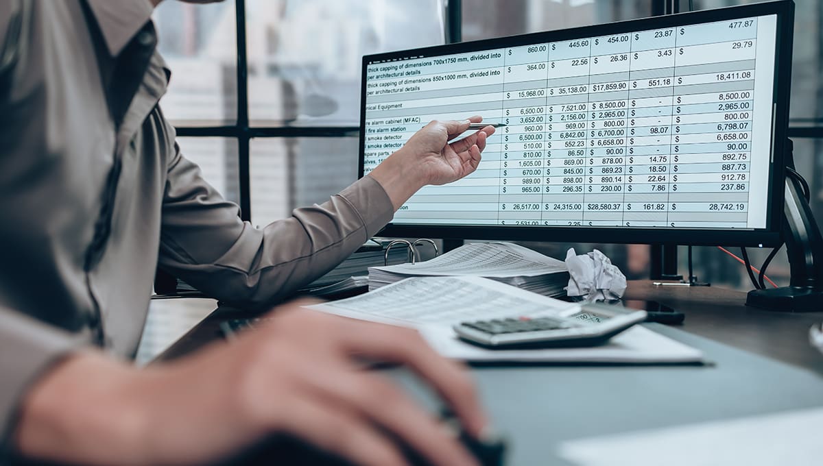 Worker at desk gesturing with pen towards budget numbers in a spreadsheet on a desktop computer monitor