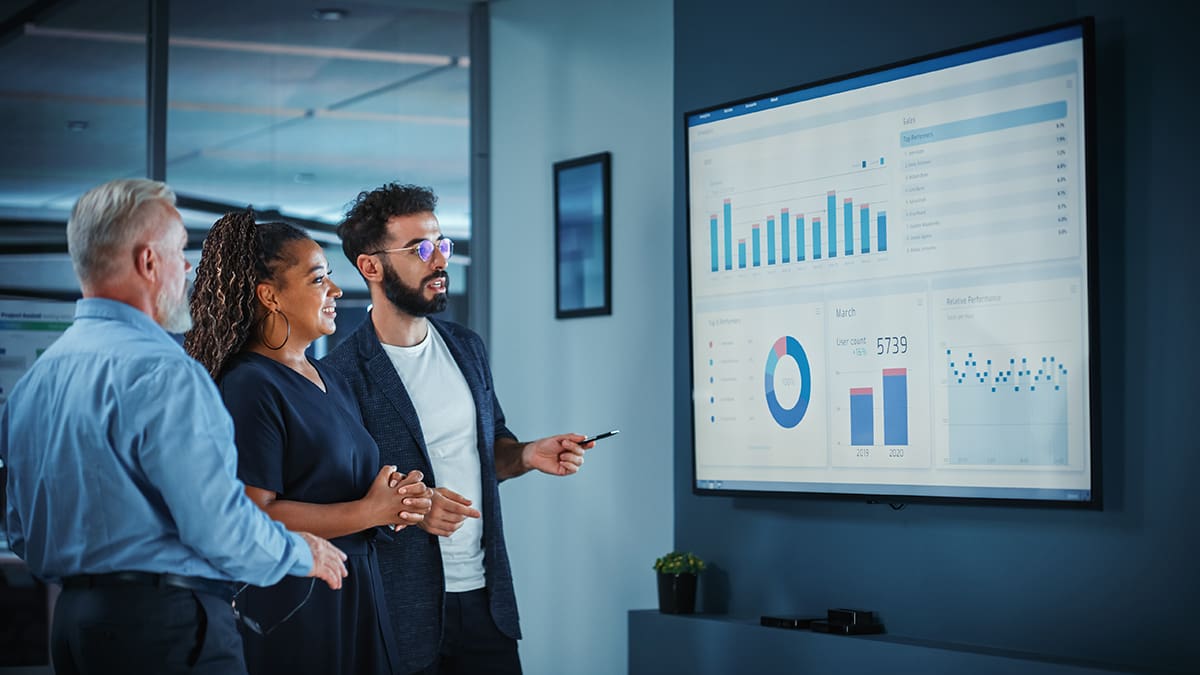Office team discusses digital marketing results in front of large monitor in conference room